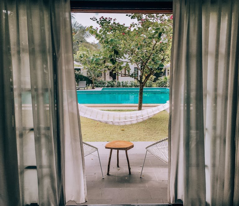 Charming poolside view through curtains with hammock and seating, ideal for a summer vacation.