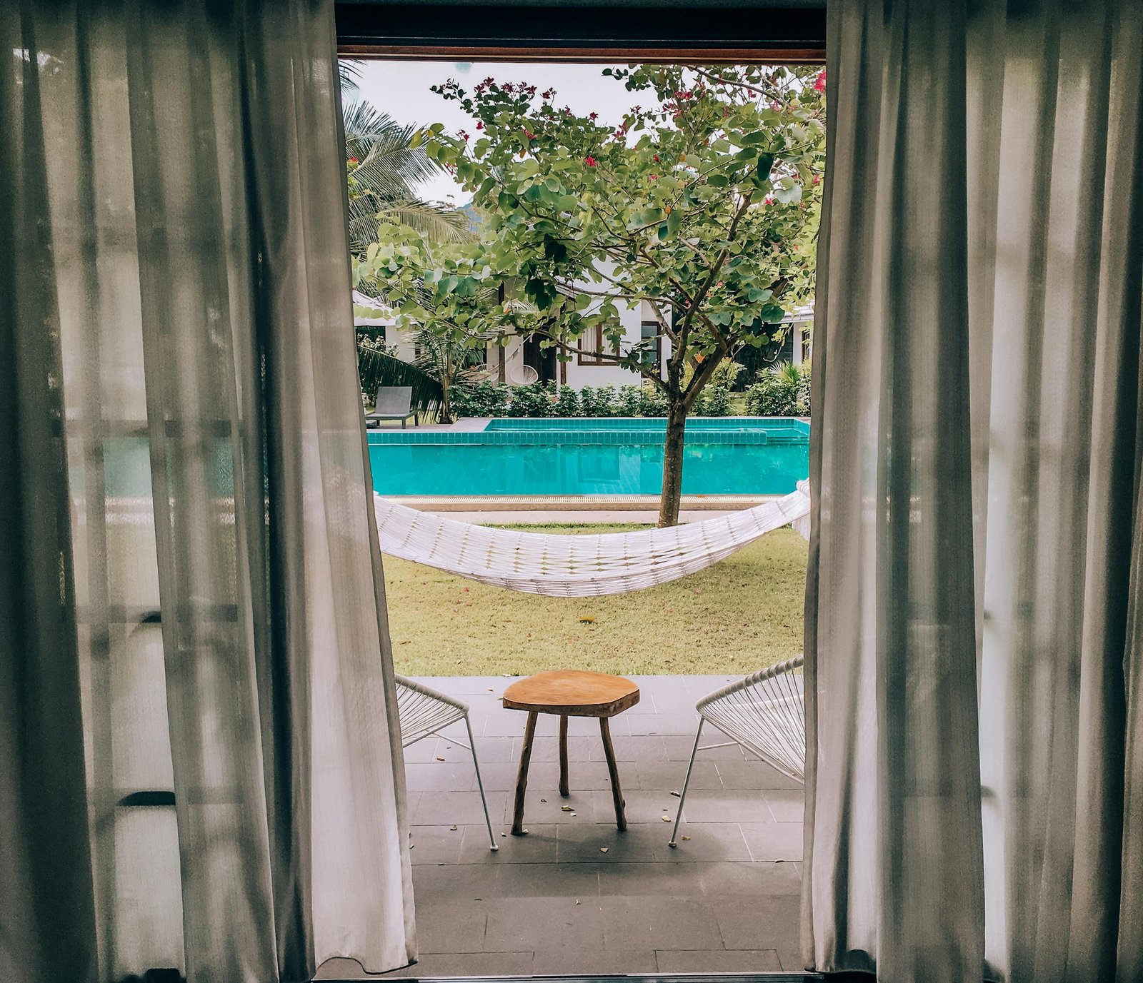 Charming poolside view through curtains with hammock and seating, ideal for a summer vacation.