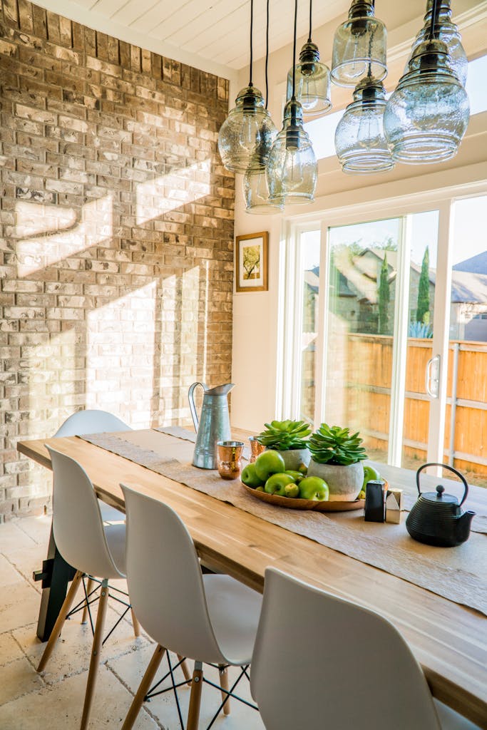 5 Tren Ruang Makan 2026 yang Akan Mengubah Tampilan Rumahmu 3 Sunlit dining room with contemporary decor and a rustic brick wall, featuring stylish lighting and a wooden table.