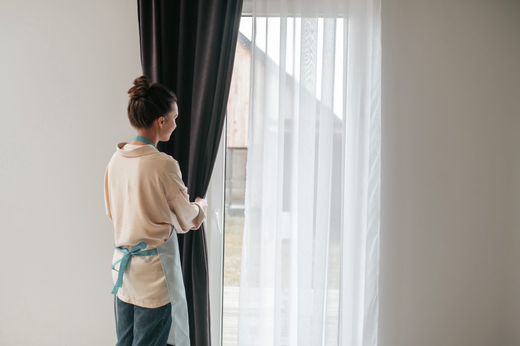 7 Manfaat Blackout Curtain yang Baik Bagi Rumahmu dan Keluargamu 8 young woman standing near widnow fixing curtains