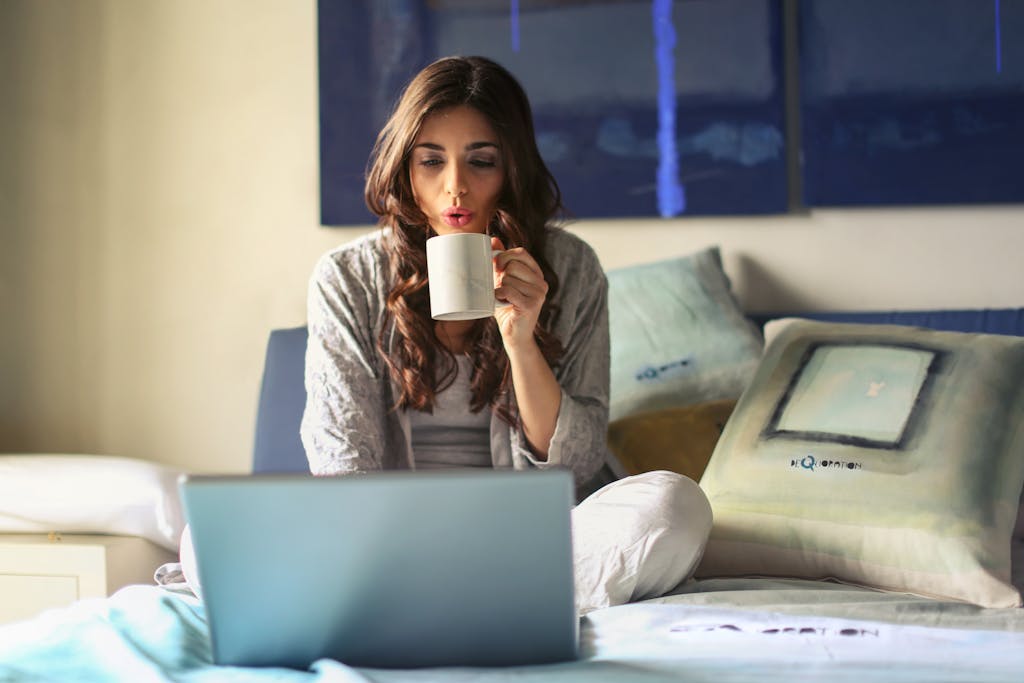 Cara Memilih Kursi Kerja untuk WFH 2 A woman enjoying coffee while working from home in a cozy bedroom setting with a laptop.